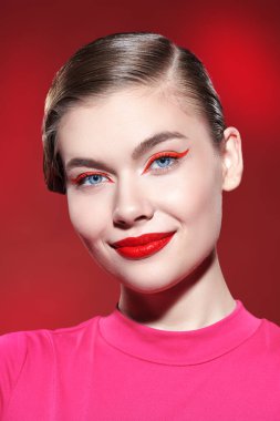 Parlak kırmızı rujlu güzel bir sarışın kız ve kırmızı arka planda büyüleyici bir şekilde gülümseyen kırmızı okları var. Makyaj ve makyaj malzemeleri. Güzellik konsepti.