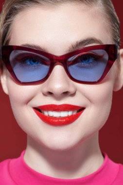 Gözlük stili. Parlak makyajlı ve zarif gözlüklü güzel bir kadın kırmızı arka planda poz veriyor. Makyaj ve makyaj malzemeleri. Yakın çekim..
