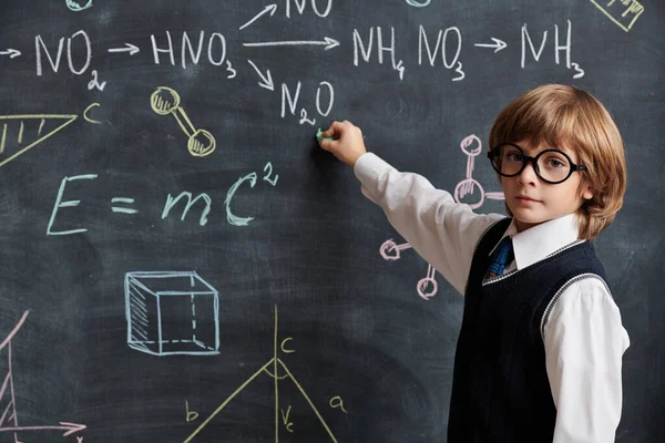 Eğitim. Düzgün okul üniforması ve gözlükleri olan zeki bir çocuk tahtaya oturmuş kimyasal formüller ve şemalar yazıyor. Akıllı çocuklar..  