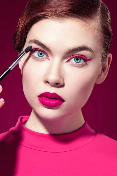Parlak pembe rujlu ve gözlerinde pembe oklar olan güzel bir manken kız koyu kırmızı arka planda bir fırçayla göz farı sürüyor. Güzellik, makyaj ve kozmetik. Makyaj araçları.