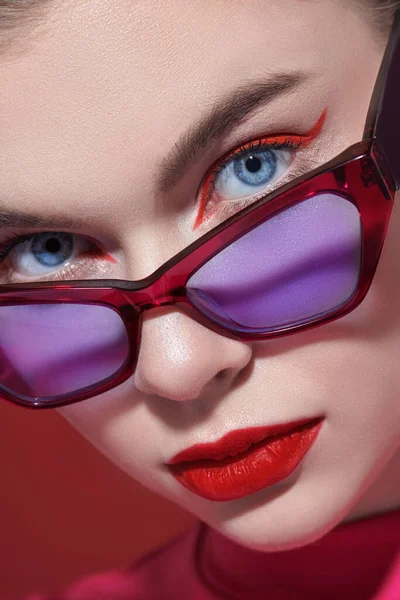 Parlak makyajlı ve zarif gözlüklü genç bir kadın kırmızı arka planda poz veriyor. Makyaj ve makyaj malzemeleri. Gözlük stili. Yakın çekim..