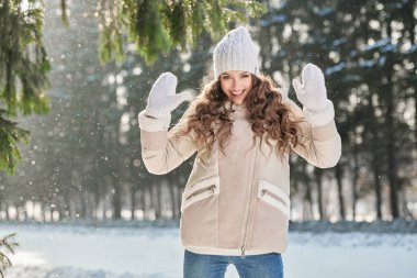 Sıcak kışlık ceketli şık bir kız, örgü şapkalı ve eldivenli bir kız, güneşli bir kış gününde parkta sevinir. Kış modası. İyi ruh hali, duygular.