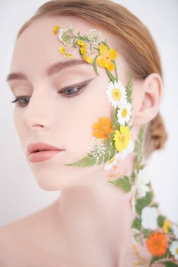 Çekici kızıl saçlı, zarif makyajlı ve yüzünde ve omzunda yaz çiçekleri olan bir kızın yakın çekimi. Beyaz arka plan. Kozmetik ve parfüm. Yazın kokusu..