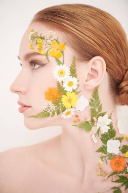 Güzel, kızıl saçlı, zarif makyajlı ve yüzünde yaz çiçekleri olan genç bir kadın başka tarafa bakıyor. Beyaz arka plan. Parfüm. Makyaj ve makyaj malzemeleri. Güzellik ve çiçekler.