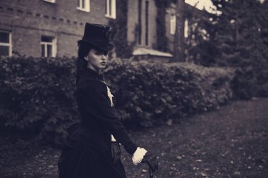 19. yüzyılın katı siyah takım elbiseli, kendinden emin ve sofistike bir bayan yeşil yapraklarla çevrili tuğla bir evde yürüyor. Uzayı kopyala Siyah ve beyaz çekim.