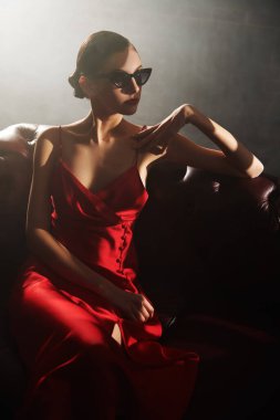 Akşam kıyafetleri ve aksesuarları. Zarif siyah gözlüklü ve kırmızı saten elbiseli güzel bir kız dumanla dolu karanlık bir çatı katında deri bir kanepede oturuyor. Güzellik ve moda.