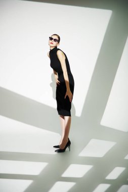 Stüdyoda gölgeli beyaz arka planda poz veren zarif siyah elbiseli ve şık güneş gözlüklü sevimli genç bayan. Tam boy çekim. 