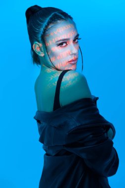 Parlak akşam makyajı ve parlak neon yazıtları olan esmer bir kız. Yüzüne yansıyor. Üstü ve deri ceketiyle mavi bir arka planda poz veriyor. Güzellik, makyaj ve kozmetik konsepti. 