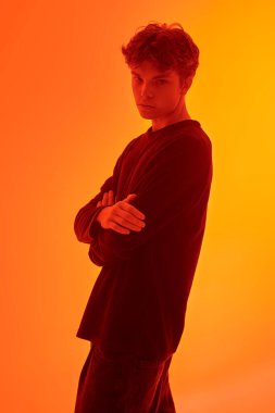 Dalgalı siyah saçlı, kolları çapraz duran depresif yakışıklı bir adamın portresi. Turuncu bir arka planda stüdyo çekimi. Gençler ve akıl sağlığı.