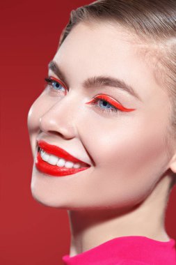 Parlak kırmızı rujlu güzel bir sarışın kız ve kırmızı arka planda büyüleyici bir şekilde gülümseyen kırmızı okları var. Makyaj ve makyaj malzemeleri. Güzellik konsepti.
