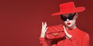 Zarif giyimli, parlak makyajlı ve siyah güneş gözlüklü genç bir kadın şık kırmızı deri bir çanta tutuyor. Kırmızı arka plan. Moda ve güzellik. Deri aksesuarlar.