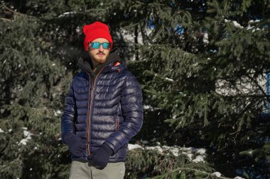 Kışlık ceketli yakışıklı bir esmer adam portresi örgü bir şapka ve güneş gözlüğü kış parkında karla kaplı köknar ağaçlarının yanında duruyordu. Kış modası.