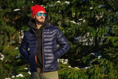 Kışlık ceketli, kendine güveni tam bir esmer adam, örgü bir şapka ve güneş gözlüğü kış parkında karla kaplı köknar ağaçlarının yanında duruyor. Kış modası.