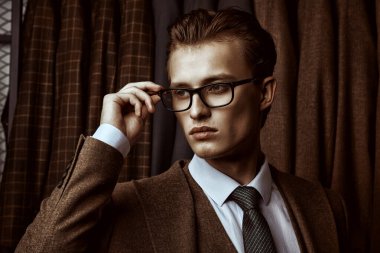 Zarif kahverengi takım elbiseli yakışıklı sarışın bir adam ve lüks klasik takım elbiseli bir mağazanın arka planında poz veren gözlüklü. Erkeklerin iş modası.