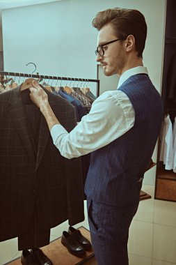 Zarif giyimli, gözlüklü bir esmer şık erkek giyim mağazasından pahalı bir takım elbise seçer. Erkek işi modası. Yan plan çekimi.