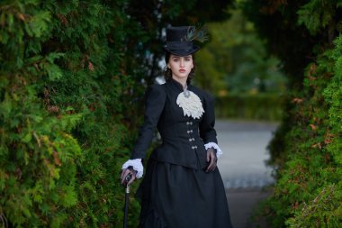 19. yüzyılın sonları ve 20. yüzyılın başlarına ait tarihi yeniden yapılanma. Katı siyah elbiseli ve şapkalı zarif, koyu renk saçlı bir bayan parkta yürüyor. Tarihsel makyaj ve saç stili.