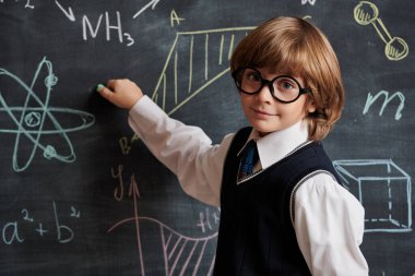 Eğitim. Düzgün okul üniforması ve gözlükleri olan zeki bir çocuk tahtaya oturmuş kimyasal formüller ve şemalar yazıyor. Akıllı çocuklar..  