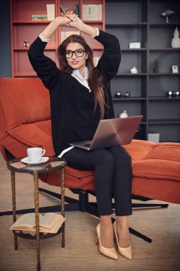 Zarif, güzel bir iş kadını bir kanepede oturup dizüstü bilgisayarla çalışırken kollarını uzatıyor. Modern iç mekan. İş ve iletişim. 