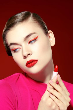 Güzellik kızı. Makyajda kırmızı renk. Parlak makyajlı güzel bir kız modelin portresi. Elinde muhteşem kırmızı bir ruj tüpü var. Burgundy arkaplanı. 