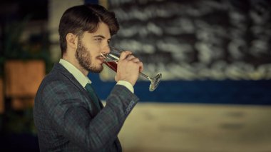 Zarif klasik takım elbiseli, kendine güvenen yakışıklı bir esmer adam lüks dairelerde dikilip bir bardak şarap içiyor. Erkek güzelliği, moda. İş stili. İşadamı. 