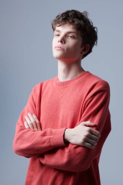 Gri arka planda kolları çapraz duran kırmızı kazaklı yakışıklı bir adamın portresi. Erkeklerin gençlik modası. 