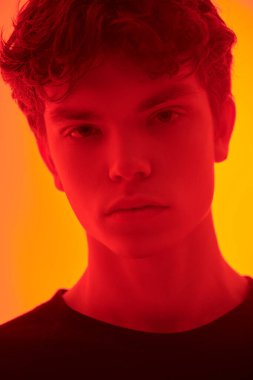 Kırmızı ışıklandırmalı, turuncu arka planda duran kapkara saçlı yakışıklı bir adamın stüdyo fotoğrafı. İnsanlar, duygular. 