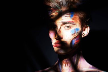 Karanlığın dışına bakan yakışıklı, boyalı yüzlü bir adamın sanat portresi. Yüzüne bir ışık huzmesi düşer. Kopyalama alanı olan siyah arkaplan. Vücut boyası.