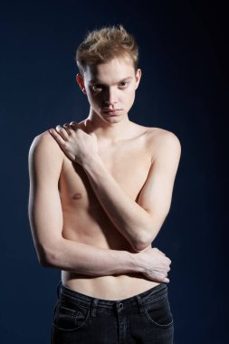 Erkek güzeli. Koyu mavi arka planda çıplak gövdeli siyah kotla poz veren yakışıklı bir genç adam portresi. Stüdyo çekimi.