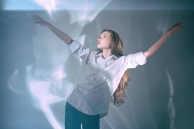 Beyaz gömlekli, uzun kahverengi saçlı güzel bir kız gölgelere bakar ve kollarını açar. Işık ve gölgeler arasında stüdyo çekimi.