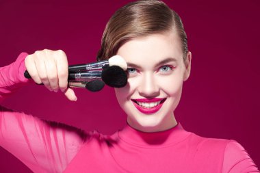 Parlak pembe rujuyla gülümseyen güzel bir sarışın kız ve fuşya arka planında çeşitli makyaj fırçaları tutan pembe okları var. Makyaj ve makyaj malzemeleri. Makyaj araçları.
