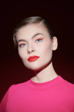 Güzellik konsepti. Parlak kırmızı rujlu ve gözlerinde kırmızı oklar olan çekici sarışın bir kız koyu bir arka planda poz veriyor. Makyaj ve kozmetik. 