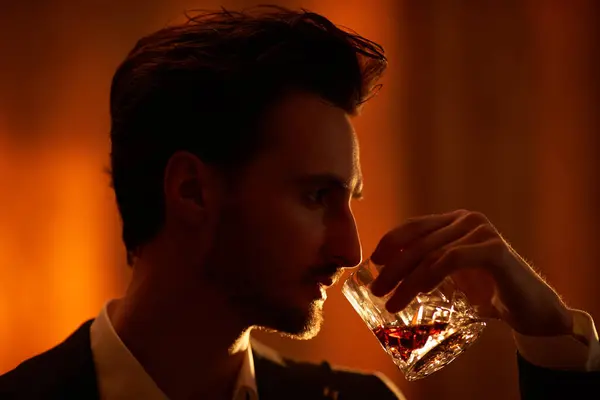 Sideview shot of handsome brunet man standing in a room with dark lighting and drinking whiskey. Men\'s beauty, lifestyle. Noir style.