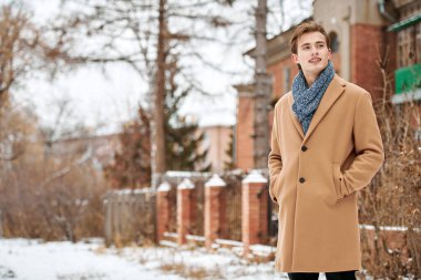 Pardösü ve atkısı olan neşeli yakışıklı genç adam şehrin caddesinde bir tuğla evin yanında duruyor ve gülümsüyor. Erkek modası ve yaşam tarzı.