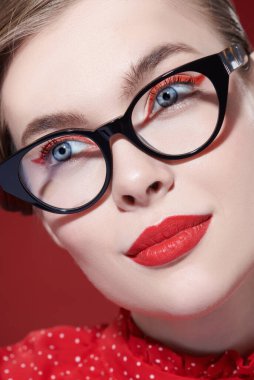Gözlük stili. Parlak makyajlı ve zarif gözlüklü güzel bir kadın kırmızı arka planda poz veriyor. Makyaj ve makyaj malzemeleri. Yakın çekim..