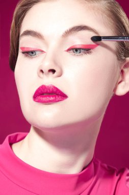 Parlak pembe rujlu ve gözlerinde pembe oklar olan güzel bir model kız bir fırçayla göz farı sürüyor. Fuchsia geçmişi. Güzellik, makyaj ve kozmetik. Makyaj araçları.