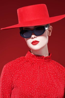 Moda ve güzellik. Parlak makyajlı, kırmızı elbiseli, kırmızı şapkalı ve güneş gözlüklü çekici bir model kız kırmızı arka planda poz veriyor. Makyaj ve kozmetik. 