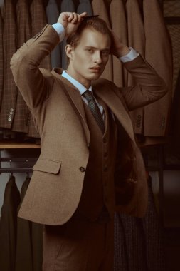 Zarif kahverengi takım elbiseli yakışıklı bir sarışının portresi premium erkek giyim mağazasında duruyor ve saçını tarıyor. Erkeklerin iş modası.