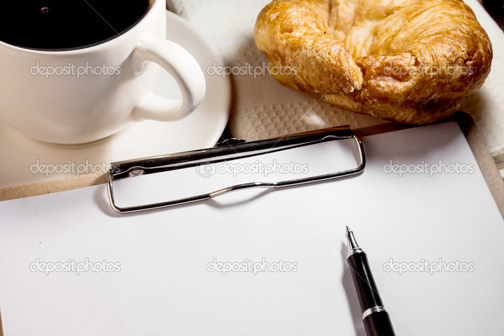 blank white notebook,pen,cup of coffee and croissant on the de