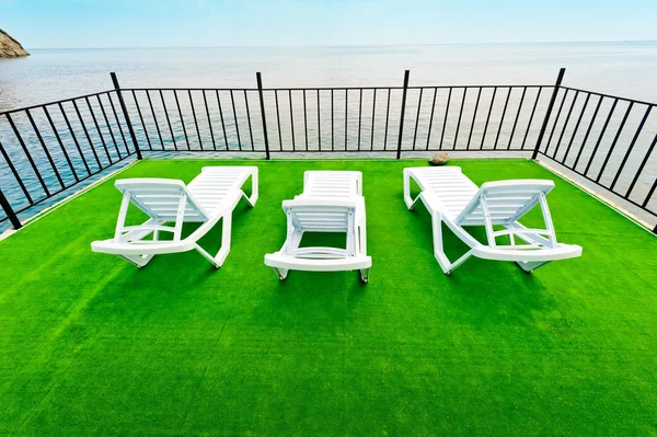 Trois chaises longues de plage blanches sur un pont vert avec vue sur la mer . — Photo