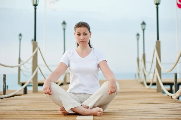 Schönes Mädchen in Lotusposition, das sich auf dem Pier am Meer ausruht — Stockfoto