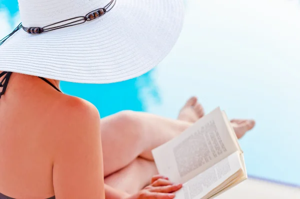 Fille dans un chapeau lisant un livre près de la piscine, assis dans une chaise longue — Photo