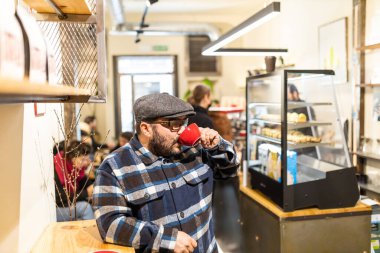 Ekose gömlekli ve yün şapkalı sakallı bir adam kahve dükkanında espresso tadıyor.