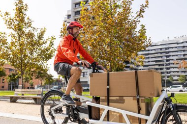 young courier with red clothing and helmet riding cargo bike , riding along the city bike path on his way to deliver a package. clipart