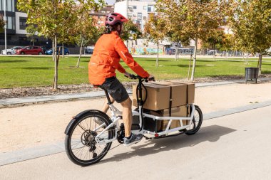 Kırmızı elbiseli ve kasklı genç bir kurye kargo bisikleti sürüyor. Bisiklet yolunda bir paket teslim etmeye gidiyor..