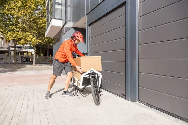 young courier with red clothing and helmet riding cargo bike arriving at the shipping destination to deliver a package to a city address