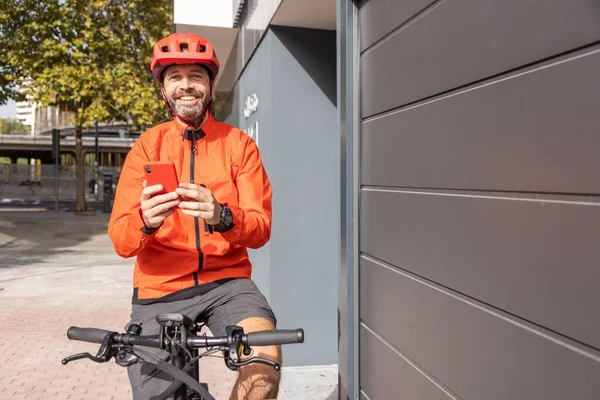 young courier with red clothing and helmet riding cargo bike arriving at the shipping destination to deliver a package to a city address, checking the shipping address on your smart phone and smiling