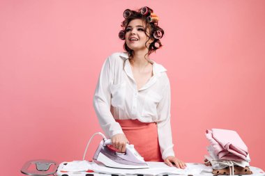 Gülümseyen ve ütü masasında ütü yapan zarif elbiseli bir bayan. Güzel bir kadın pembe arka planda izole edilmiş.