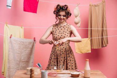 Gülümseyen, güzel esmer ev hanımı pembe bir duvarda elbiseleri olan iplerden oluşan bir arka planda aşçılık yapıyor. Kadın yumurta kırıyor, mutfak aletleri ayakta duruyor..