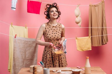 Gülen, puantiyeli bir elbise içinde neşeli bir kız, yumurta çırpıyor, kıyafetlerle bir halatın arkasında yemek pişiriyor. Pembe arka planda saçları kıvırcık saçlı bir ev kadını..