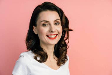 Gülümseyen, pembe duvar arkasında poz veren genç bir kadın. Pembe arka planda kırmızı dudakları olan çekici bir esmer..